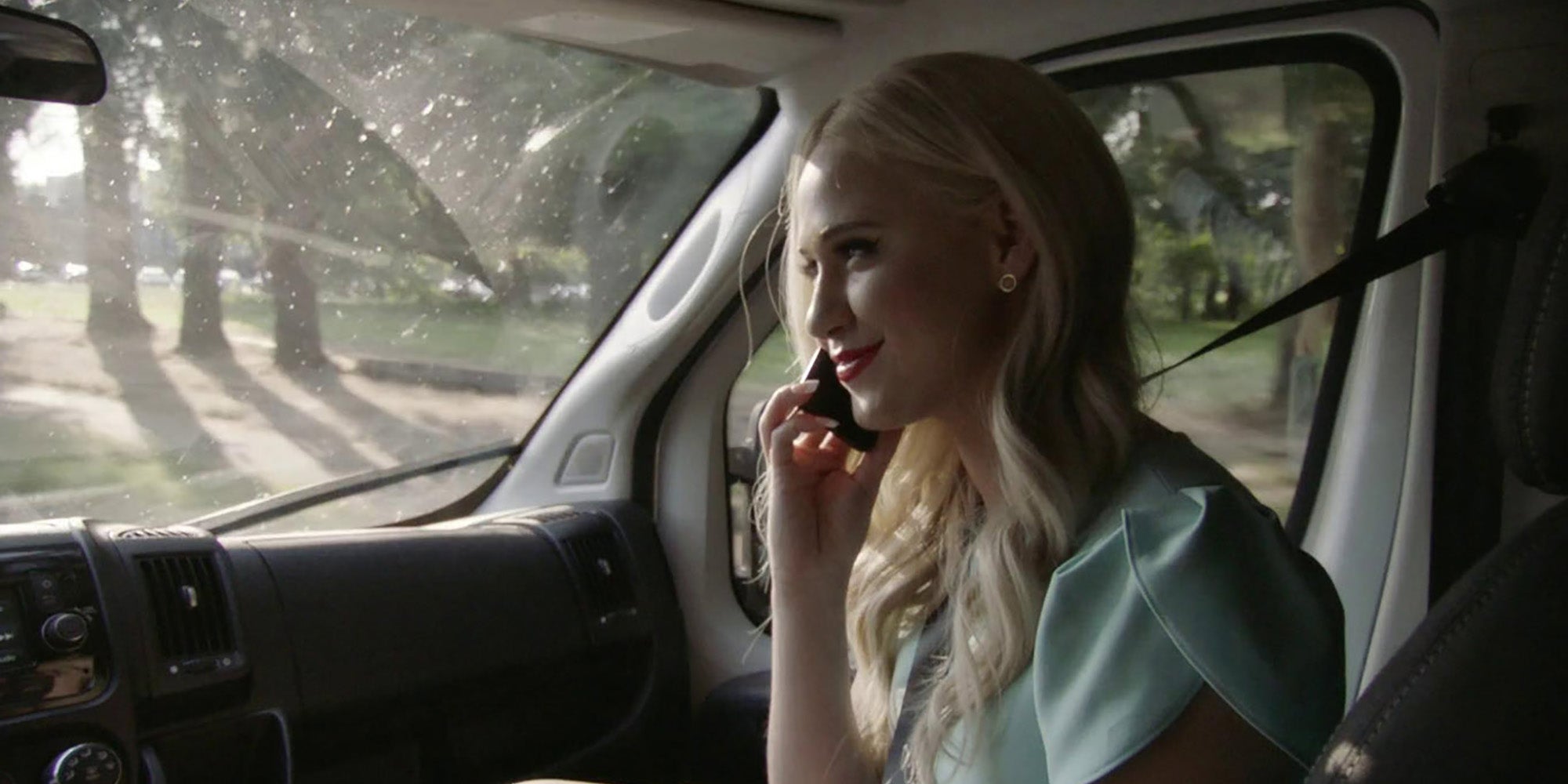 A person with blonde hair riding in the passenger seat of a car and listening to a phone. Maria Bakalova in Borat Subsequent Moviefilm