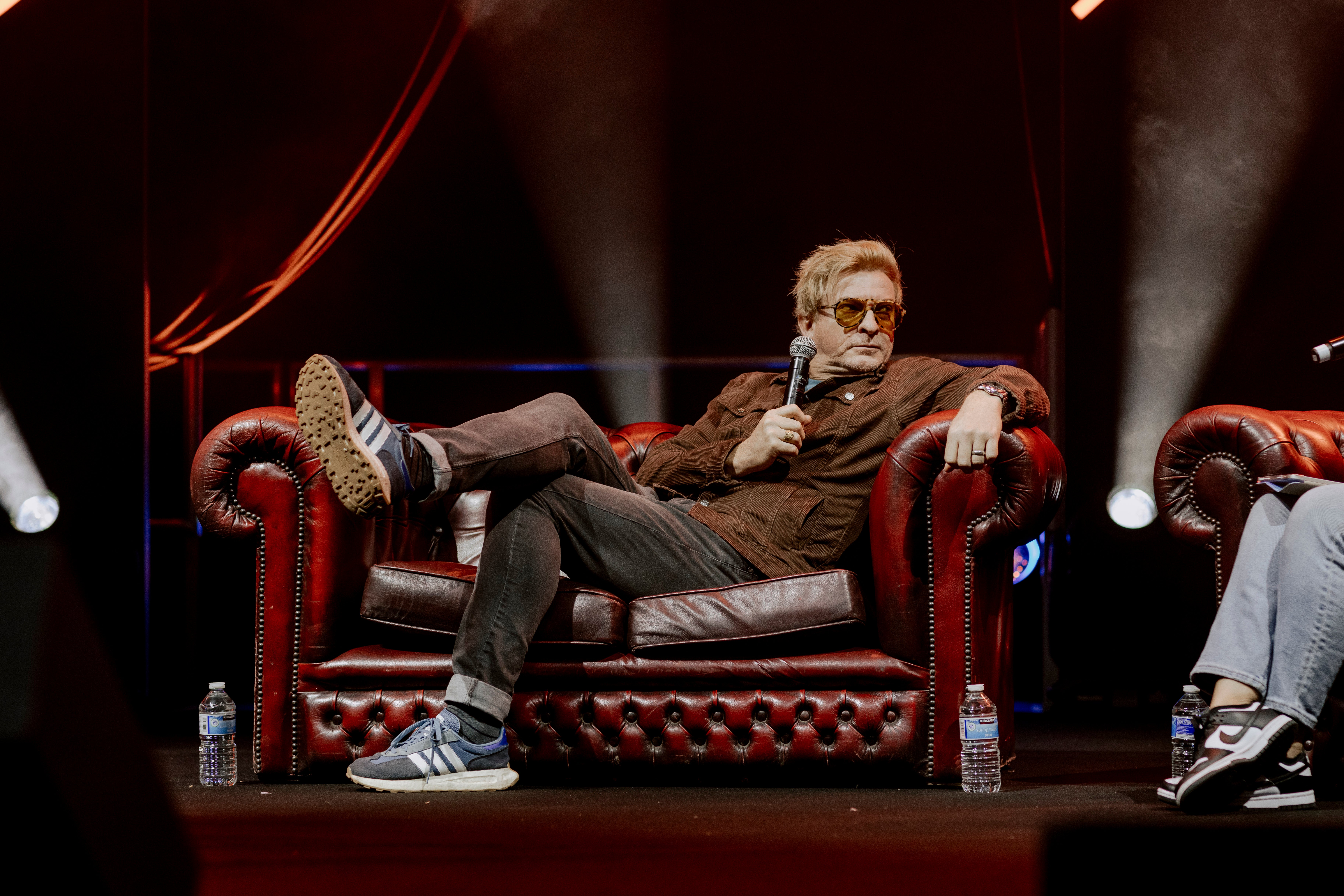 Photograph of Rhys Darby reclining on a couch onstage at MCM